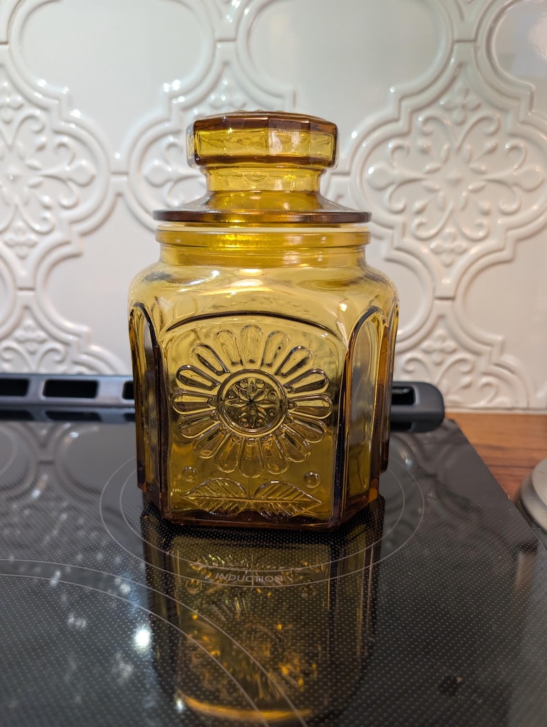 May include: Amber-colored glass jar with a lid. The jar has a hexagonal shape with a floral design on the front. The lid is also amber-colored and has a geometric design. The jar is sitting on a black surface.