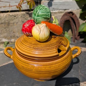 May include: A ceramic soup tureen with a lid, featuring a decorative arrangement of vegetables. The tureen is a warm, earthy brown with a textured, striped design. The lid is adorned with a cabbage, tomato, onion, and carrot.