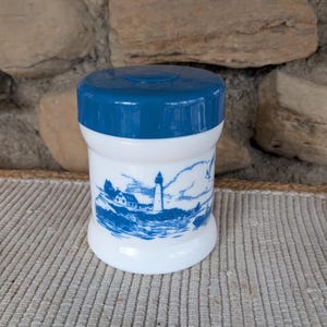 Puede incluir: Un tarro de cerámica blanco y azul con tapa azul. El tarro presenta una ilustración azul de un faro, edificios y el océano. El tarro es cilíndrico y se asienta sobre una superficie texturizada.