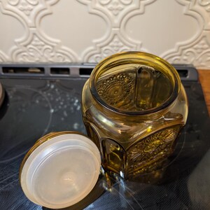 May include: Amber-colored glass jar with a decorative lid. The jar has a hexagonal shape with embossed floral patterns. The lid is off to the side, revealing the jar's interior. The jar is sitting on a black surface.