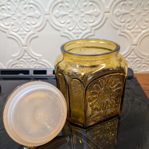 May include: Amber-colored glass jar with a decorative sunflower design and a clear plastic lid. The jar is square with rounded corners. The lid is off to the side. The jar is sitting on a black surface.