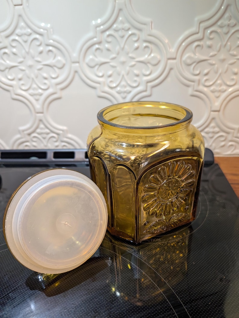 May include: Amber-colored glass jar with a decorative sunflower design and a clear plastic lid. The jar is square with rounded corners. The lid is off to the side. The jar is sitting on a black surface.