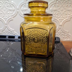 May include: Amber-colored glass jar with a lid. The jar has a square shape with rounded corners and a decorative embossed floral design on the front. The lid is also amber-colored and fits snugly on top.