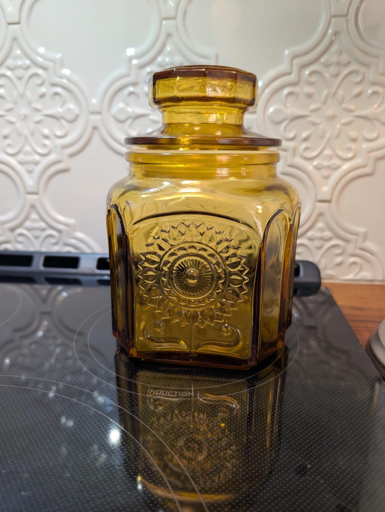 May include: Amber-colored glass jar with a lid. The jar has a square shape with rounded corners and a decorative embossed floral design on the front. The lid is also amber-colored and fits snugly on top.