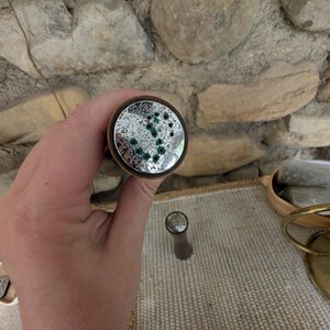 Op de afbeelding: Een decoratieve flesstopper met een cirkelvormige, spiegelende bovenkant met groene accenten. De stopper heeft een bronzen metalen rand en een bruine steel. Het ontwerp doet denken aan een melkwegstelsel of een abstract patroon. De stopper is ontworpen voor wijnflessen.
