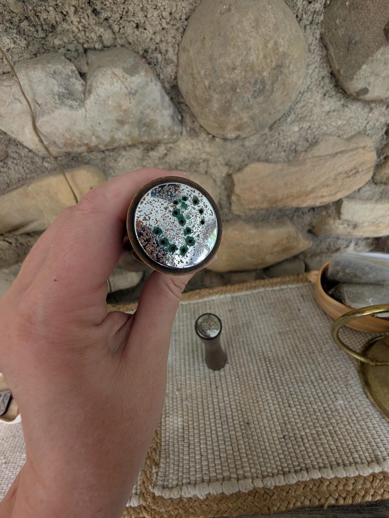 Op de afbeelding: Een decoratieve flesstopper met een cirkelvormige, spiegelende bovenkant met groene accenten. De stopper heeft een bronzen metalen rand en een bruine steel. Het ontwerp doet denken aan een melkwegstelsel of een abstract patroon. De stopper is ontworpen voor wijnflessen.
