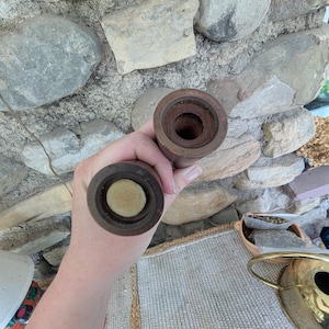 Op de afbeelding: Twee houten pepermolens, elk met een donkerbruine afwerking. De bovenkant van &eacute;&eacute;n molen is open en onthult het interieur. De bovenkant van de andere molen heeft een lichtgekleurd, cirkelvormig stuk. De molens worden tegen een stenen muur gehouden.