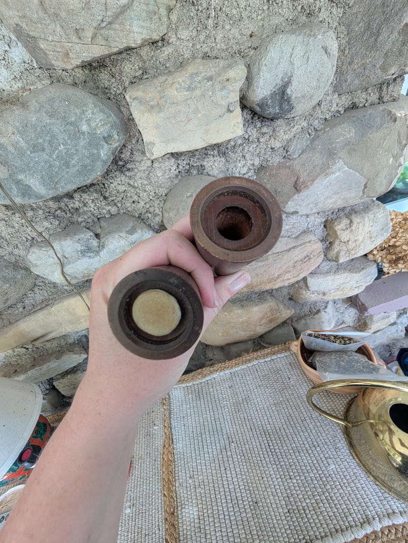 Op de afbeelding: Twee houten pepermolens, elk met een donkerbruine afwerking. De bovenkant van &eacute;&eacute;n molen is open en onthult het interieur. De bovenkant van de andere molen heeft een lichtgekleurd, cirkelvormig stuk. De molens worden tegen een stenen muur gehouden.