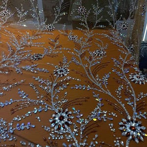 以下が含まれることがあります： 繊細な花柄刺繍が施されたシアー生地のクローズアップ。銀色と虹色のクリスタルが植物模様に配置され、茶色と赤色のアクセントが施されています。生地は温かみのあるオレンジ色の表面にドレープされています。