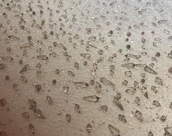 May include: Close-up of a beige fabric with a pattern of small, clear crystals. The fabric is covered in a dense layer of crystals, creating a shimmering effect. The text "100% HAND MADE" is visible in the lower right corner of the image.