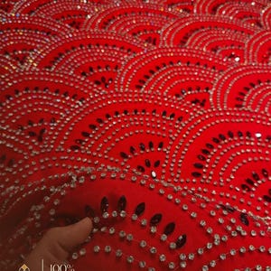 Op de afbeelding: Close-up van een felrode stof versierd met een herhalend, golfachtig patroon van sprankelende strass-steentjes en donkere, druppelvormige versieringen. De woorden "100% HAND MADE" zijn zichtbaar. De stof wordt op zijn plaats gehouden door een hand.