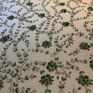 Könnte beinhalten: Weißer Stoff mit einem aufwendigen Blumenmuster. Das Muster zeigt silberfarbene Stiele und Blätter, verziert mit grünen und klaren kristallartigen Elementen. Das Design ist gleichmäßig über den Stoff verteilt.