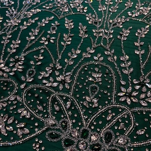 May include: Emerald green fabric with intricate silver and crystal embellishments. The design features floral and botanical patterns, with sparkling crystals and silver beading. The fabric has a luxurious, textured appearance, suitable for formal wear or decorative projects.