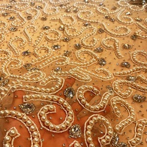 May include: Beige mesh fabric with a delicate pattern of white pearls and clear rhinestones. The fabric is embellished with a repeating design of small circles and swirls.