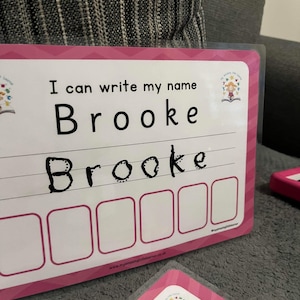 Pode incluir: Cart&atilde;o de aprendizagem rosa e branco com o texto "Eu posso escrever meu nome Brooke" e um espa&ccedil;o para escrever o nome. O nome Brooke est&aacute; escrito em marcador preto abaixo do texto. H&aacute; seis espa&ccedil;os em branco abaixo do nome para praticar a escrita.