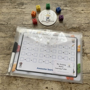 May include: A clear plastic folder containing a white dry-erase board. The board has a grid of squares with plus and minus signs. The folder is labelled "My Amazing Maths Learning Folder".