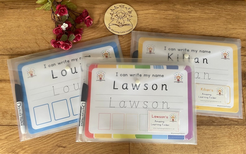 May include: Three clear plastic folders with colorful borders and white writing surfaces. Each folder has a different name printed on it: "Loui", "Lawson", and "Kilian". The folders are designed for children to practice writing their names.