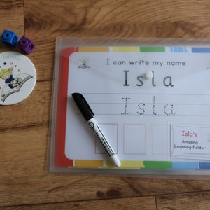 May include: A white dry-erase board with a colorful border and a clear plastic sleeve. The board has the text "I can write my name Isla" and a dotted line for practicing writing the name Isla. A black dry-erase marker is next to the board.
