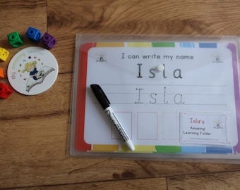 May include: A white dry-erase board with a colorful border and a clear plastic sleeve. The board has the text "I can write my name Isla" and a dotted line for practicing writing the name Isla. A black dry-erase marker is next to the board.