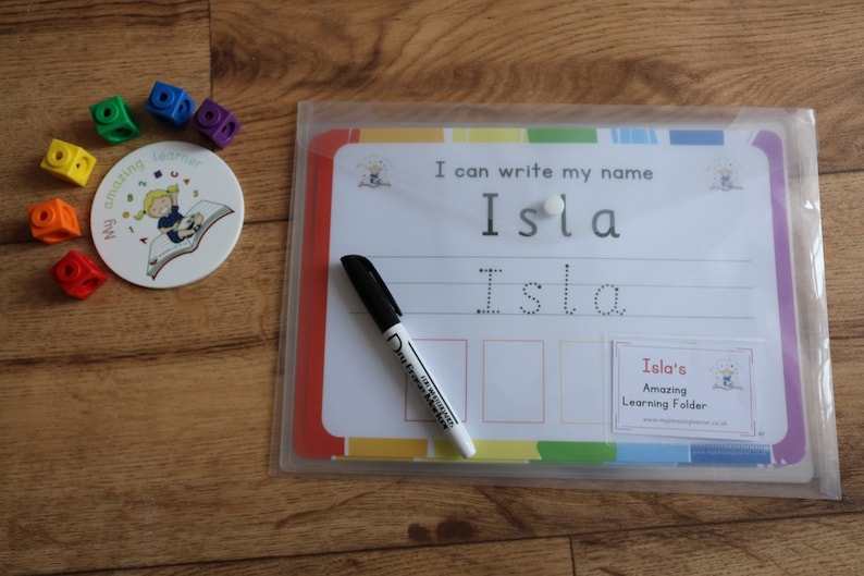 May include: A white dry-erase board with a colorful border and a clear plastic sleeve. The board has the text "I can write my name Isla" and a dotted line for practicing writing the name Isla. A black dry-erase marker is next to the board.