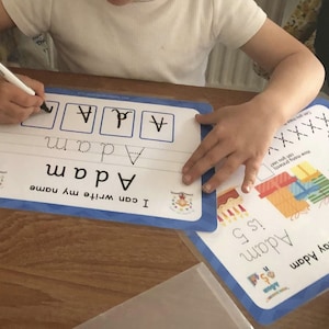 Pode incluir: Uma crian&ccedil;a est&aacute; aprendendo a escrever seu nome, "Adam", em um tapete de apagamento a seco azul e branco. O tapete tem uma se&ccedil;&atilde;o para praticar as letras do nome e uma se&ccedil;&atilde;o para escrever o nome completo. A crian&ccedil;a est&aacute; usando um marcador preto para escrever as letras.