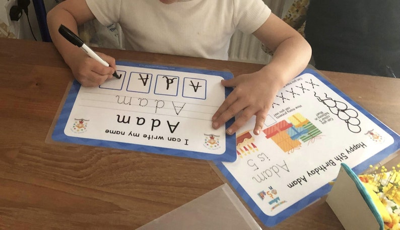 Pode incluir: Uma crian&ccedil;a est&aacute; aprendendo a escrever seu nome, "Adam", em um tapete de apagamento a seco azul e branco. O tapete tem uma se&ccedil;&atilde;o para praticar as letras do nome e uma se&ccedil;&atilde;o para escrever o nome completo. A crian&ccedil;a est&aacute; usando um marcador preto para escrever as letras.