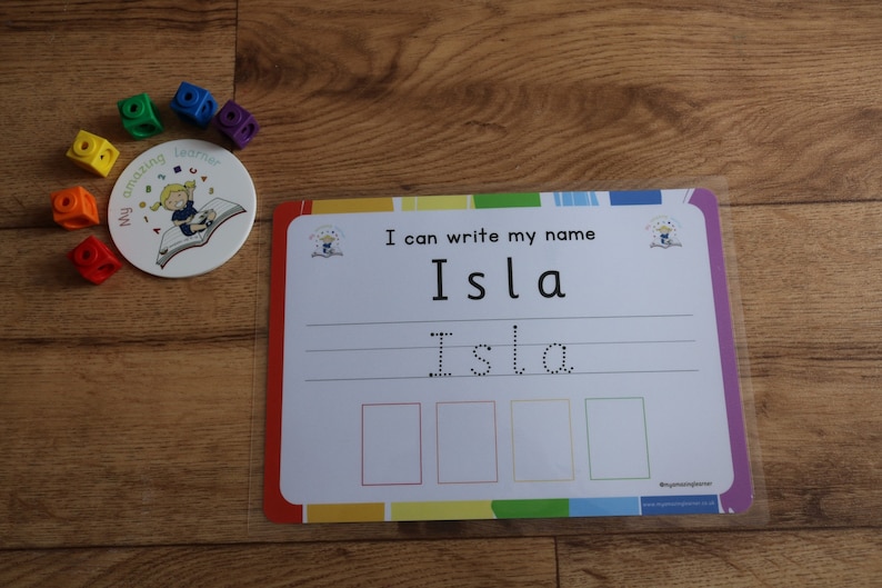 Pode incluir: Uma folha de aprendizagem laminada branca com uma borda arco-&iacute;ris. A folha tem o texto "Eu posso escrever meu nome Isla" e uma linha pontilhada para tra&ccedil;ar o nome Isla. H&aacute; tamb&eacute;m quatro caixas vazias com linhas pontilhadas para tra&ccedil;ar.