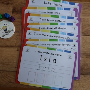 May include: A set of colorful laminated cards with different activities for kids to practice writing and tracing. The cards feature phrases like "Let's doodle", "I can trace lines", "I can trace patterns", "I can draw 2D shapes", "I can trace my numbers 1-10", "I can trace my alphabet letters", and "I can write my name". The last card has the name "Isla" written on it.