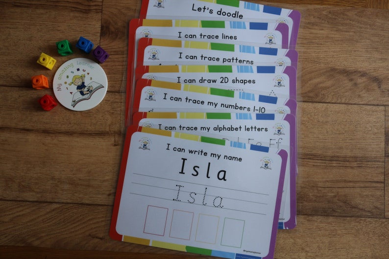May include: A set of colorful laminated cards with different activities for kids to practice writing and tracing. The cards feature phrases like "Let's doodle", "I can trace lines", "I can trace patterns", "I can draw 2D shapes", "I can trace my numbers 1-10", "I can trace my alphabet letters", and "I can write my name". The last card has the name "Isla" written on it.