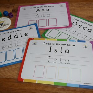 May include: Four colorful laminated cards with dotted lines for tracing letters. Each card has a different name: Ada, Oscar, Isla, and Freddie. The cards are on a wooden surface.