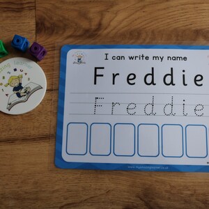 Pode incluir: Um quadro branco de apagamento a seco azul e branco com o texto "Eu posso escrever meu nome Freddie" e uma linha pontilhada para praticar a escrita do nome Freddie. H&aacute; tamb&eacute;m seis caixas vazias abaixo da linha pontilhada.
