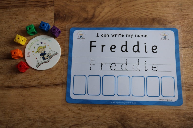 Pode incluir: Um quadro branco de apagamento a seco azul e branco com o texto "Eu posso escrever meu nome Freddie" e uma linha pontilhada para praticar a escrita do nome Freddie. H&aacute; tamb&eacute;m seis caixas vazias abaixo da linha pontilhada.