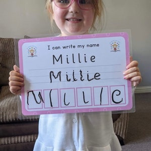 Pode incluir: Uma menina pequena com &oacute;culos rosa segura um quadro de aprendizagem rosa e branco. O quadro diz "Eu posso escrever meu nome" e tem o nome "Millie" escrito nele com marcador preto. A menina escreveu seu nome no quadro e est&aacute; praticando a escrita das letras.