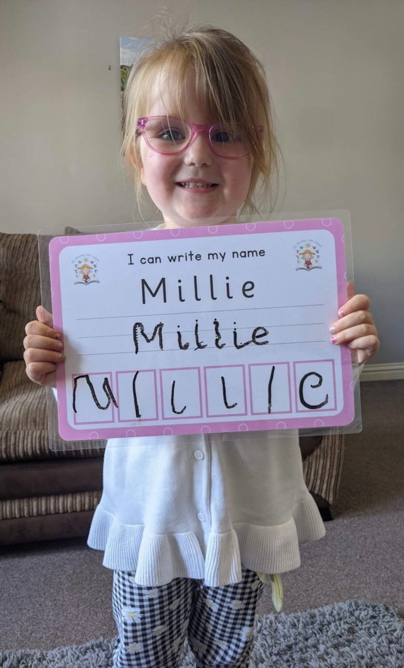 Pode incluir: Uma menina pequena com &oacute;culos rosa segura um quadro de aprendizagem rosa e branco. O quadro diz "Eu posso escrever meu nome" e tem o nome "Millie" escrito nele com marcador preto. A menina escreveu seu nome no quadro e est&aacute; praticando a escrita das letras.