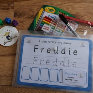 May include: A blue and white dry-erase learning folder with the text "I can write my name Freddie" and a dotted line for tracing the name. The folder includes a clear plastic pouch with dry-erase markers and a white eraser.
