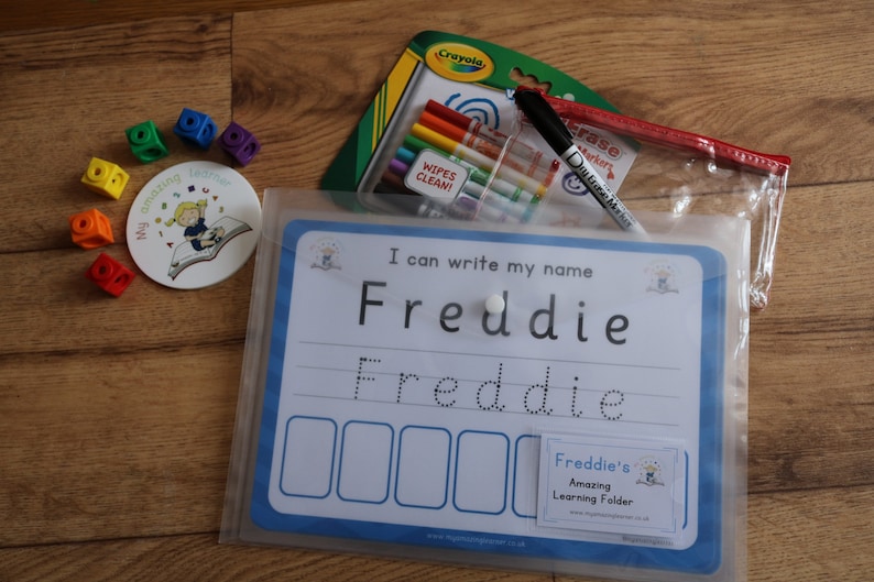 May include: A blue and white dry-erase learning folder with the text "I can write my name Freddie" and a dotted line for tracing the name. The folder includes a clear plastic pouch with dry-erase markers and a white eraser.