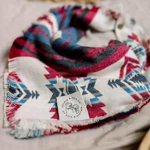 May include: A white, red, and blue patterned bandana with frayed edges. The bandana has a small white tag with a black logo that says "Archie & Fitz Co."