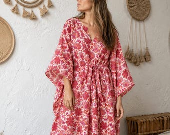 May include: A floor-length kaftan dress featuring a vibrant pink and red floral pattern on a white background. The garment has wide, loose sleeves, a V-neckline, and a drawstring waist tie that creates a gathered silhouette.