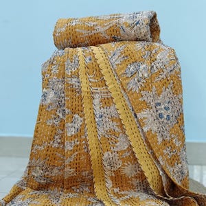 May include: A mustard yellow cotton quilt featuring a repeating floral and leaf pattern in white and grey. The fabric is detailed with visible running stitch embroidery throughout, creating a textured kantha effect across the entire surface.