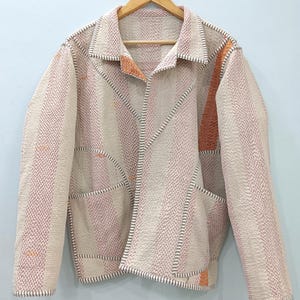 Könnte beinhalten: Eine langärmelige Jacke aus strukturiertem, gestepptem Stoff mit vertikalen Streifen in Creme, zartem Rosa und gebranntem Orange. Das Kleidungsstück verfügt über einen Kragen, eine offene Vorderseite und zwei große aufgesetzte Taschen.