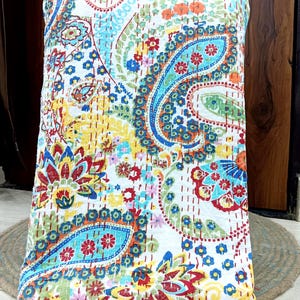 May include: A colorful paisley patterned quilt. The quilt features a white background with a vibrant design of blue, red, yellow, and orange paisley shapes and floral accents. The quilt is folded and appears to be made of soft fabric.