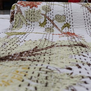 May include: A white cotton quilt featuring a repeating botanical print of brown branches and light green leaves. The fabric is decorated with rows of dark brown running stitches that create a textured, quilted appearance across the entire surface.