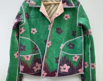 May include: A long-sleeve jacket featuring a vibrant green base with a repeating pattern of pink and dark purple flowers. The garment has a collared neckline, two front pockets, and visible white stitching along the edges and seams throughout.