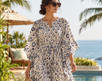 May include: A floor-length kaftan dress featuring a white base with a repeating blue floral and paisley print, a V-neckline, wide kimono-style sleeves, and a cinched waist with a drawstring tie, paired with a woven straw tote bag.