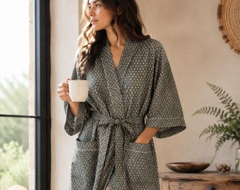 May include: A long-sleeved, floor-length robe in a muted olive green color featuring a repeating white diamond pattern. The robe includes a matching tie belt at the waist, two front pockets with white trim, and a shawl collar design.