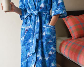 May include: A long blue robe featuring a vibrant tropical leaf pattern in shades of azure and navy. The garment includes a matching tie belt, two front pockets, and white piping trim along the lapels, cuffs, and pocket edges for a clean finish.