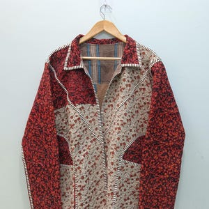 Könnte beinhalten: Eine langärmelige Jacke mit Patchwork-Design, bestehend aus abwechselnden Bahnen aus dunkelrotem, strukturiertem Stoff und cremefarbenem Stoff mit kleinem rotem Blumenmuster. Das Kleidungsstück hat einen Kragen und sichtbare weiße Nähte.