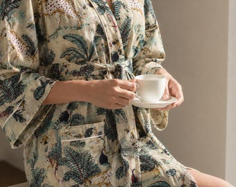 May include: A beige robe featuring a repeating pattern of cheetahs, tropical birds, and palm leaves in shades of green, black, and red. The robe has long sleeves, a tie waist, and a single front pocket with white piping trim.