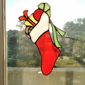 Puede incluir: Una vidriera de un calcetín navideño rojo, blanco y verde. El calcetín está relleno con una caja de regalo amarilla con un lazo naranja y un objeto marrón. La obra de arte se exhibe frente a una ventana.