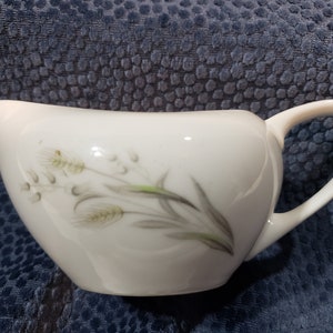 Könnte beinhalten: Ein weißer Keramik-Milchkännchen mit einem floralen Design aus Weizenähren und Blättern. Der Milchkännchen hat einen Griff und einen Ausguss.